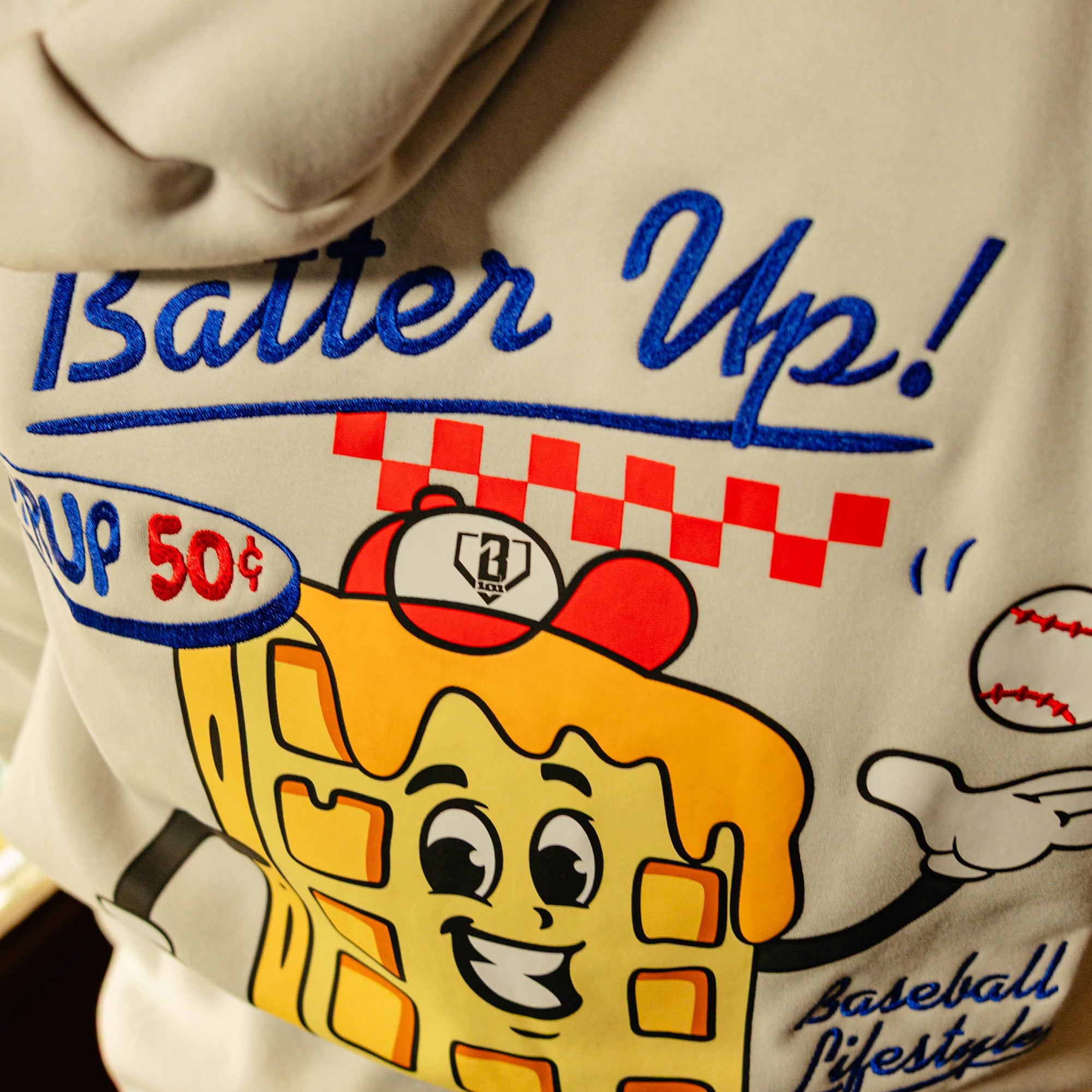 Close-up of a hoodie with a cartoon waffle character and 'Batter Up' text on a beige background