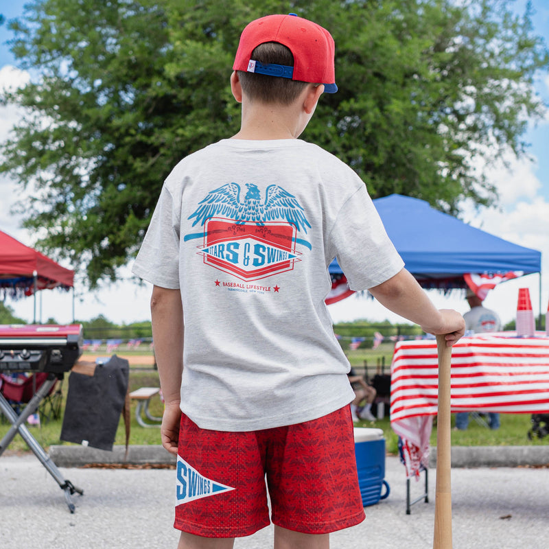 Back view of youth male model wearing red Freedom Swings shorts and heather gray Freedom Swings tee holding baseball bat at outdoor baseball event
