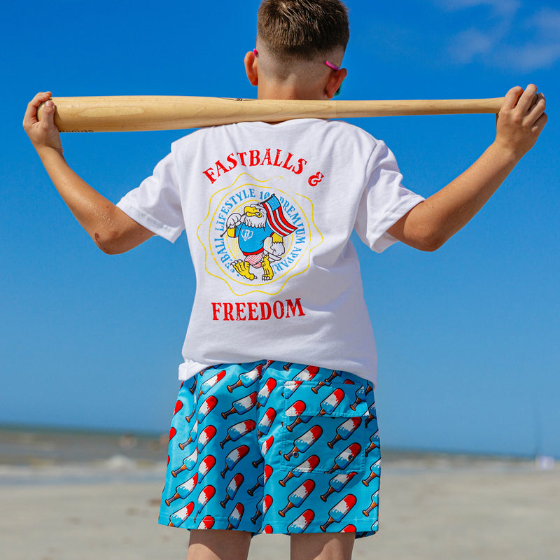 Back view of a young boy in BL101 youth popsicle bat print swim trunks and a white Fastballs & Freedom tee at the beach.