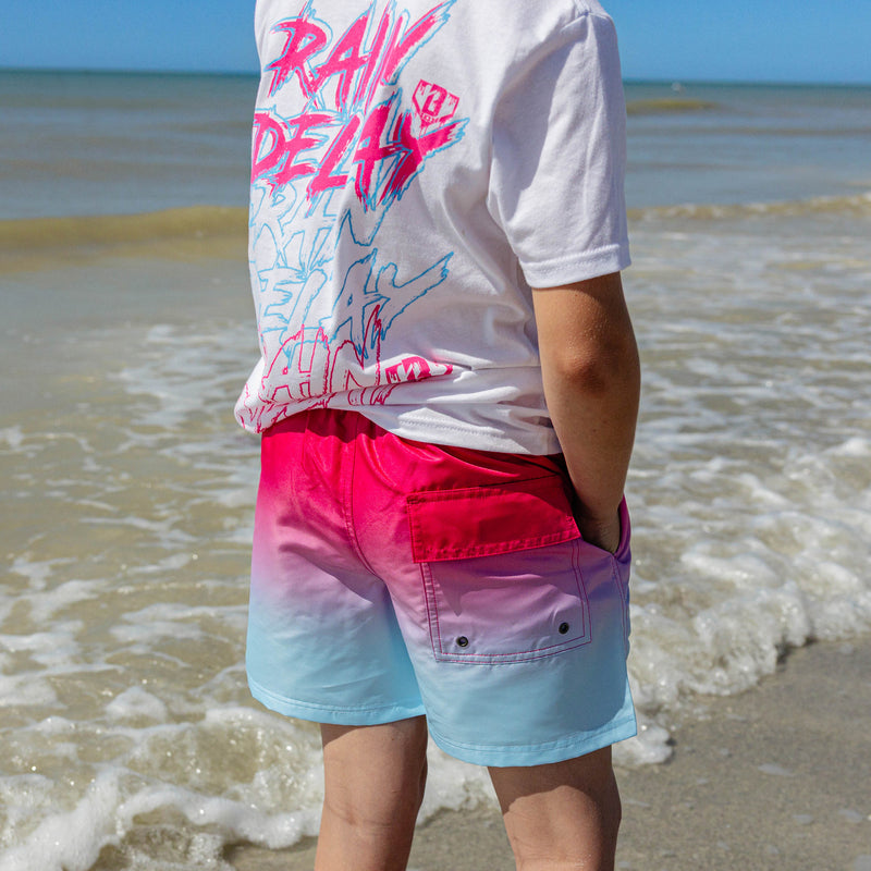 Back view of BL101 youth pink to blue ombre swim trunks showing the cargo-style snap pocket, paired with a white Rain Delay graphic tee at the water's edge.