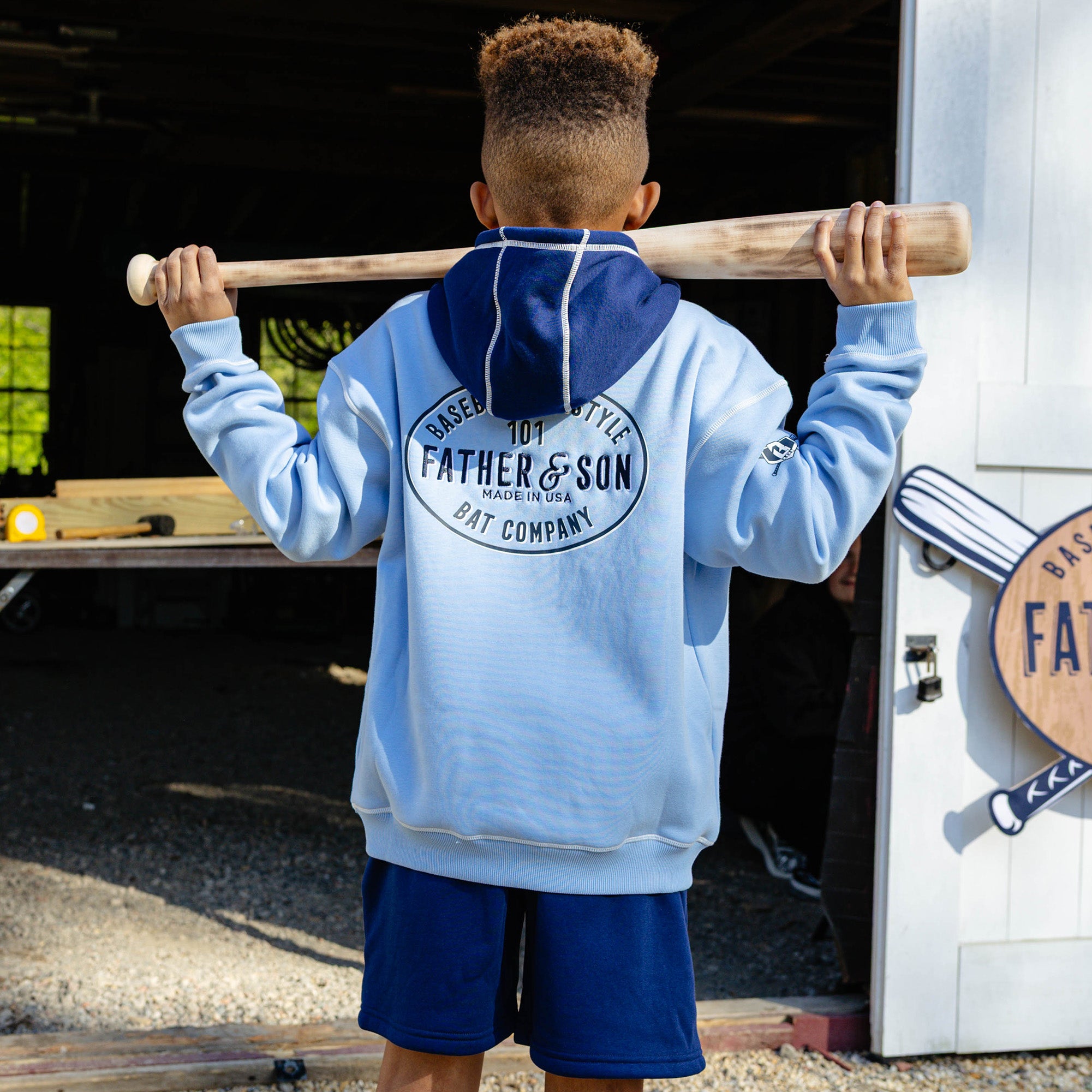 Back view of a young boy holding a wooden baseball bat across his shoulders wearing the navy youth Heritage Diamond Sweatshort paired with the columbia blue Father & Son Bat Company hoodie showing the large oval back graphic.