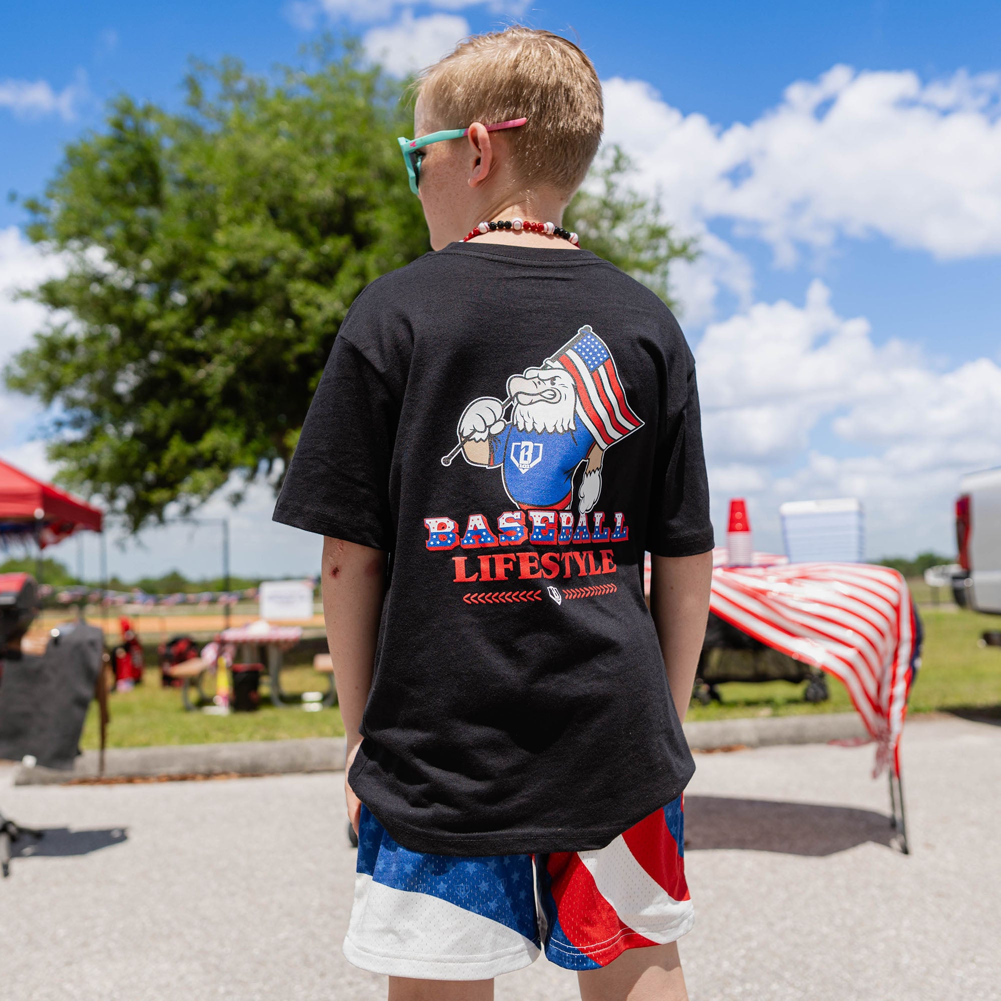Back view of youth male model wearing red white and blue Patriot Pitch shorts and black Patriot Pitch tee showing Baseball Lifestyle eagle graphic at outdoor baseball event