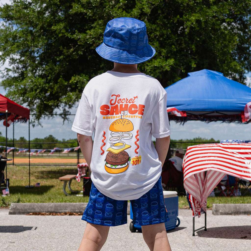 Person wearing a white secret sauce t-shirt with blue Secret Sauce burger print shorts and blue bucket hat at an outdoor event.